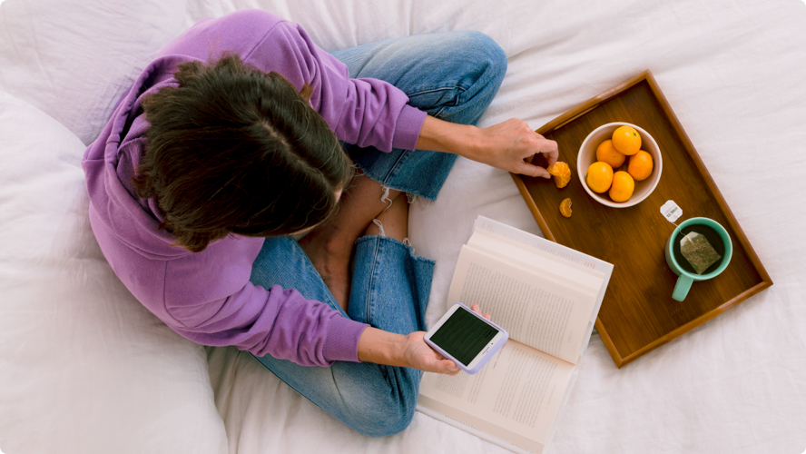 Top-down view of a person sitting cross-legged on a white comforter while browsing their phone and eating orange slices with a book open and a tray with a hot cup of tea in front of them.