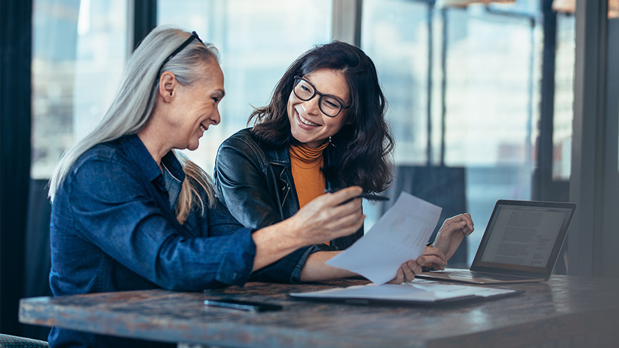 Female banker with glasses assisting an elderly women with paperwork and her laptop.