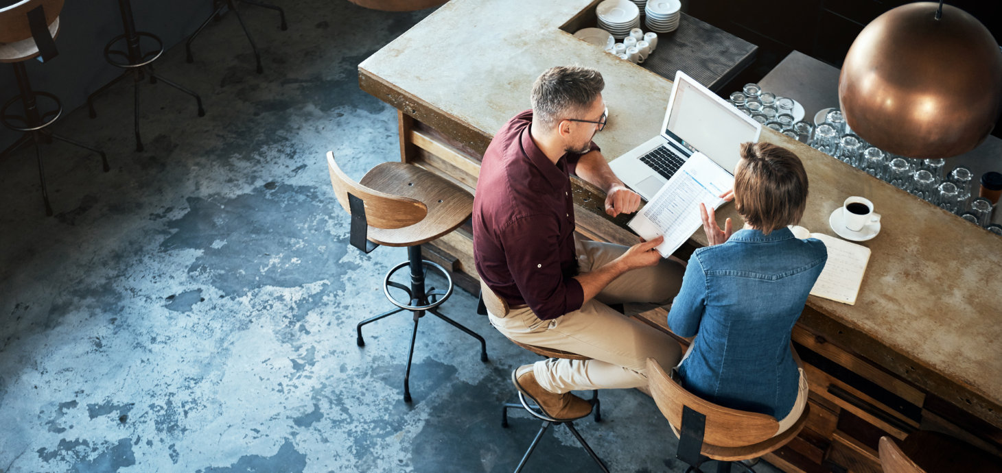 Two business people sitting at a bar top with a computer and financial paperwork.