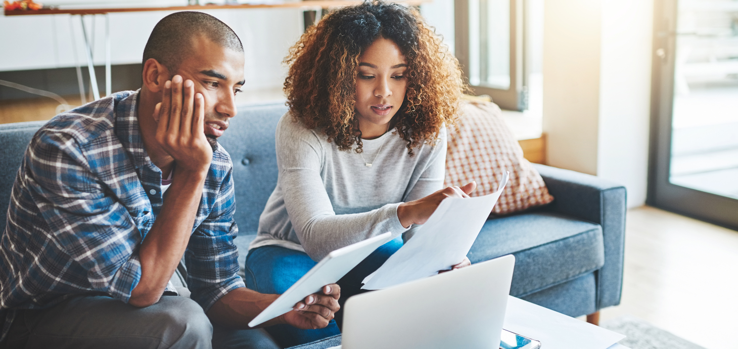 An African American male and female sitting on a couch reviewing documents and a laptop with concern.