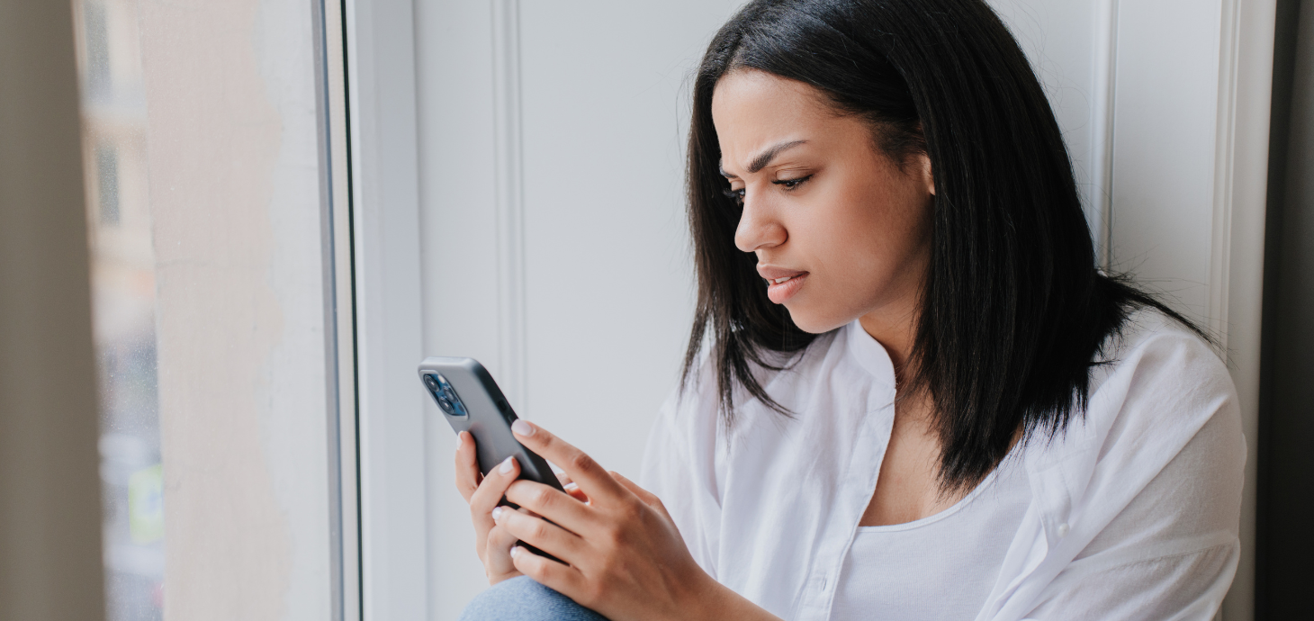 Woman sitting by a window, holding and looking at her phone with concern.