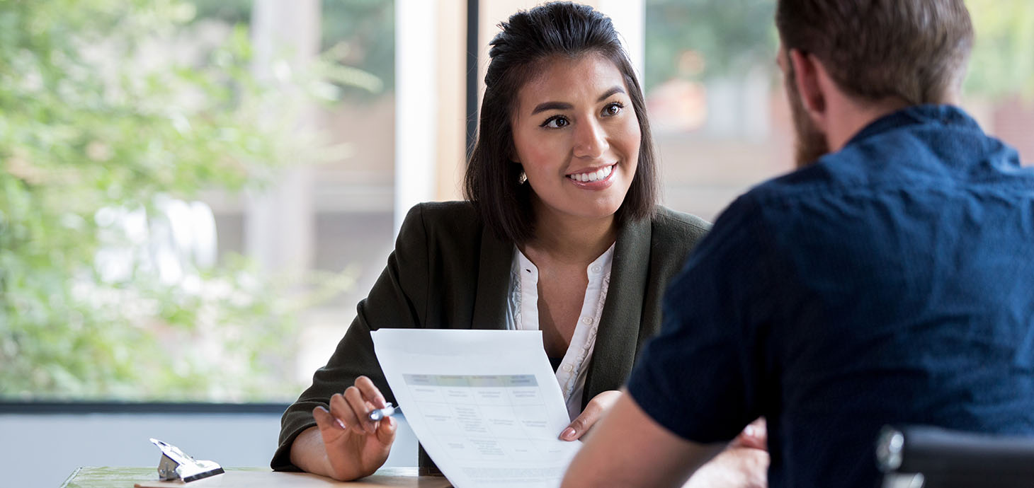 A banker, smiling as she guides a customer through a form, seated in front of a large window with trees and a building visible outside.