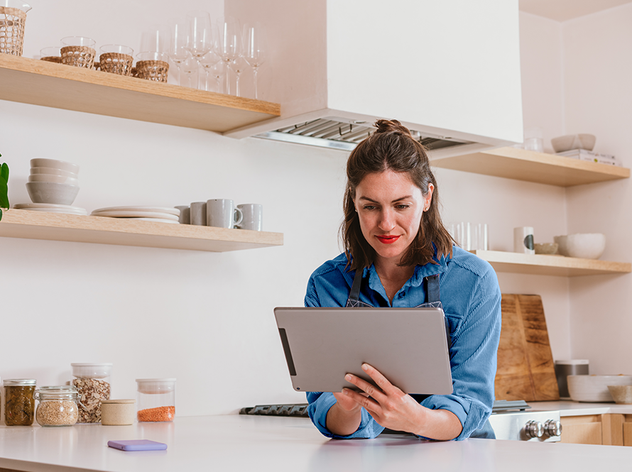 Woman standing at kitchen counter holding and looking at a tablet.